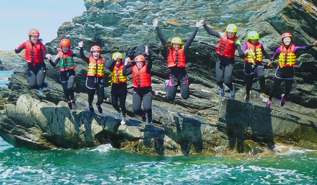 Large Coasteering group jumps together holding hands off a cliff edge